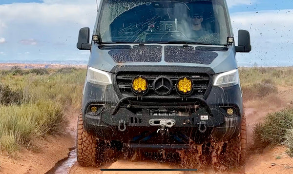 A mud-splattered Mercedes Sprinter van powers through a muddy trail. 
